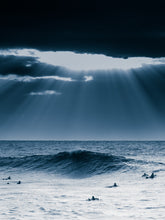 Load image into Gallery viewer, A giant wave at breaking point in the north sea, with surfers paddling around it and beams of light shining through the opening of heavy clouds. The photograph has an blue tone.
