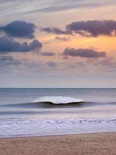 Load image into Gallery viewer, A clean wave breaks with white spray with sand from the Scheveningen beach visible. The sky has dark clouds and orange glow from the setting sun.

