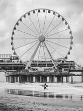Load image into Gallery viewer, The frame is filled by the ferris wheel from the pier in Scheveningen. A surfer carries a surfboard on the foreground, walking across the beach.
