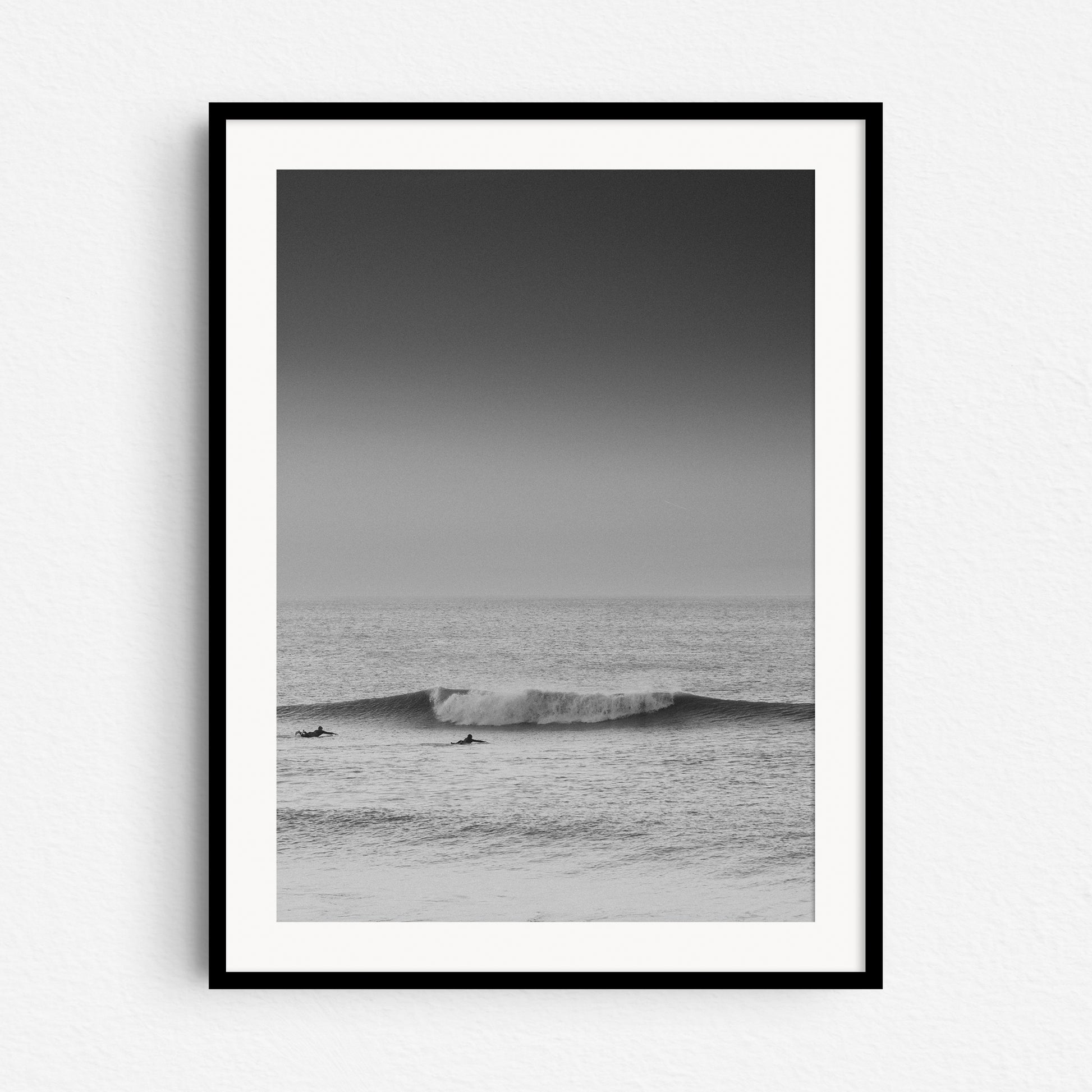 Black and white surf art from The Hague. Surfers in front of a broken wave in the North Sea. A minimalistic photo framed in black wood.