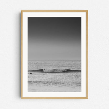 Surfers in the North Sea paddle in front of a wave. Black and white nature photography, framed in a natural wood.