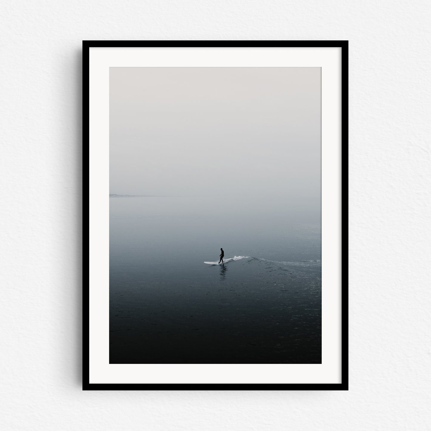 Silhouette of a surfer reflected on the water while surfing a small wave in a foggy landscape. Framed in black wood, capturing the peaceful North Sea Surf Photos essence.