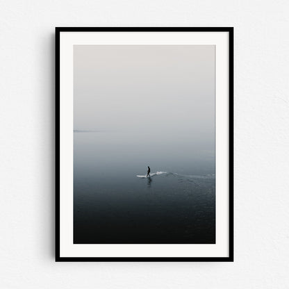 Silhouette of a surfer reflected on the water while surfing a small wave in a foggy landscape. Framed in black wood, capturing the peaceful North Sea Surf Photos essence.