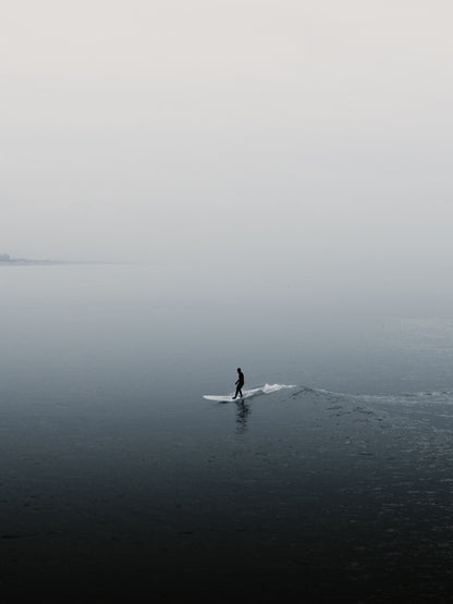 A peaceful silhouette of a surfer in Scheveningen. The landscape foggy with a shallow depth of field. Perfect for adding calming wall art with sea and surf to your space. Captured by Jop Hermans