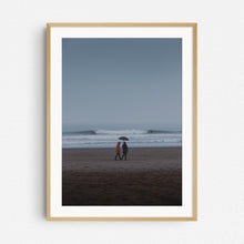 Load image into Gallery viewer, A couple holds an umbrella in a rainy, blueish surf landscape in Scheveningen. Photo print framed in natural wood.
