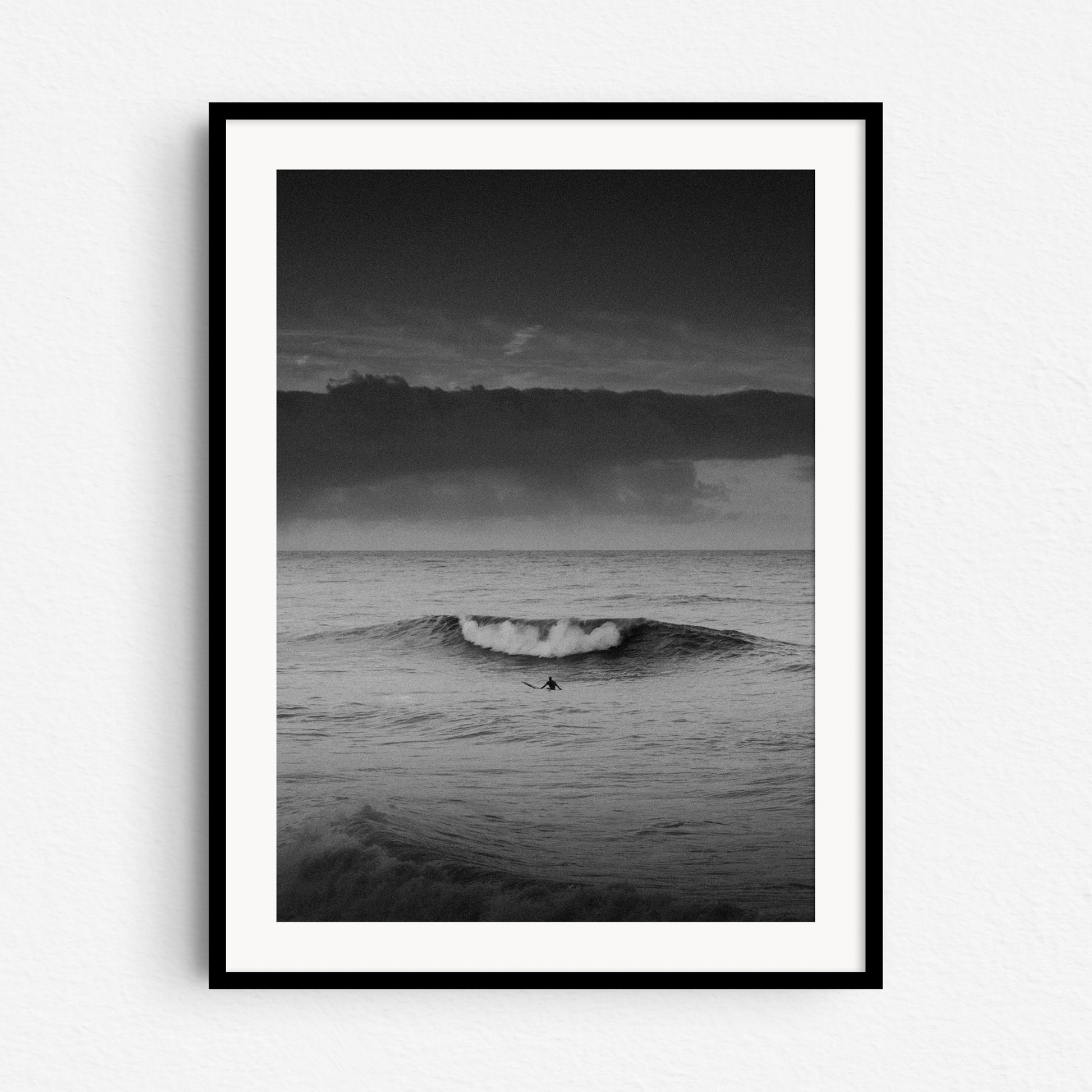 A surfer watches a wave break in front of him. A black and white surf print in a black wooden frame.