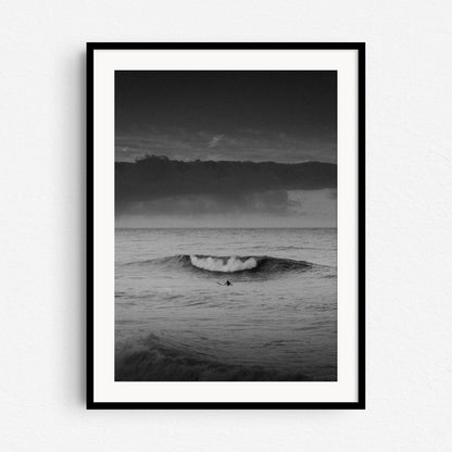 A surfer watches a wave break in front of him. A black and white surf print in a black wooden frame.