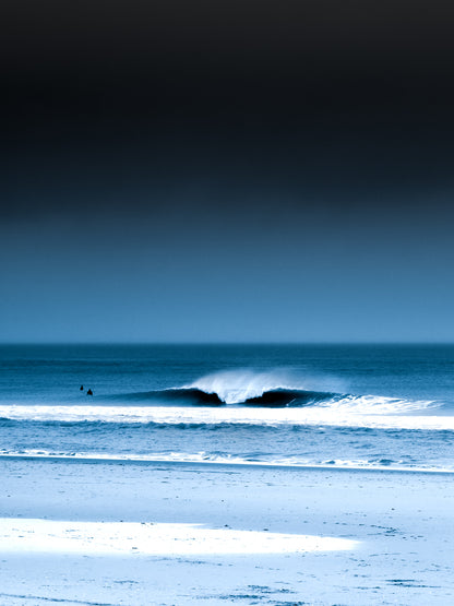 A blue photograph with two surfers in the North sea next to a breaking wave.  Minimalist coastal artwork perfect for your home.