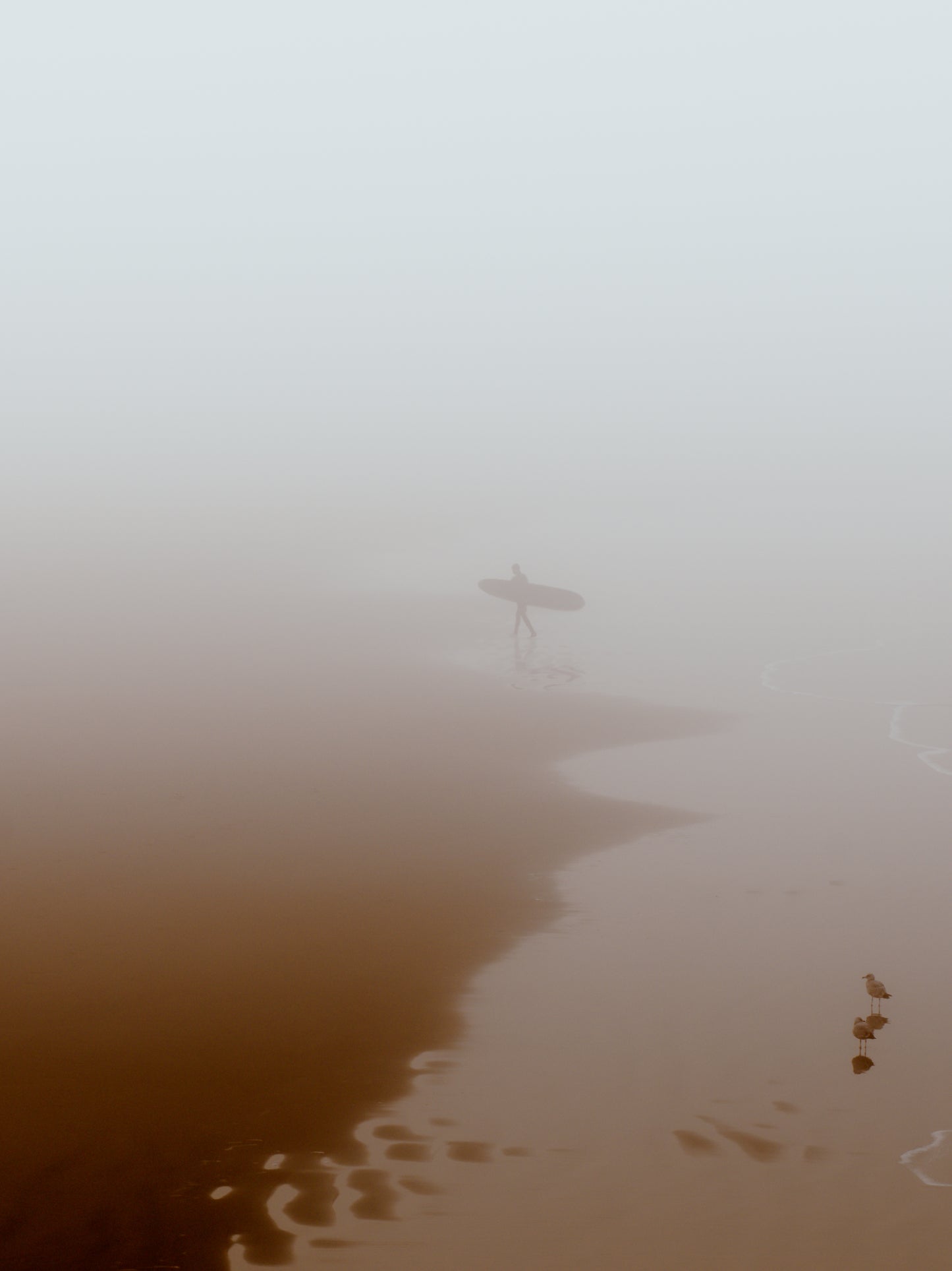 Quiet beach with a surfer leaving the water, capturing a peaceful moment in thick fog. Perfect for wall art.