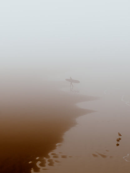 Quiet beach with a surfer leaving the water, capturing a peaceful moment in thick fog. Perfect for wall art.