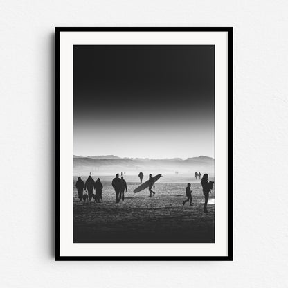 Black and white photo of a man with a longboard on a crowded Dutch beach, framed in black wood, ideal for Surf Photography Netherlands fans.