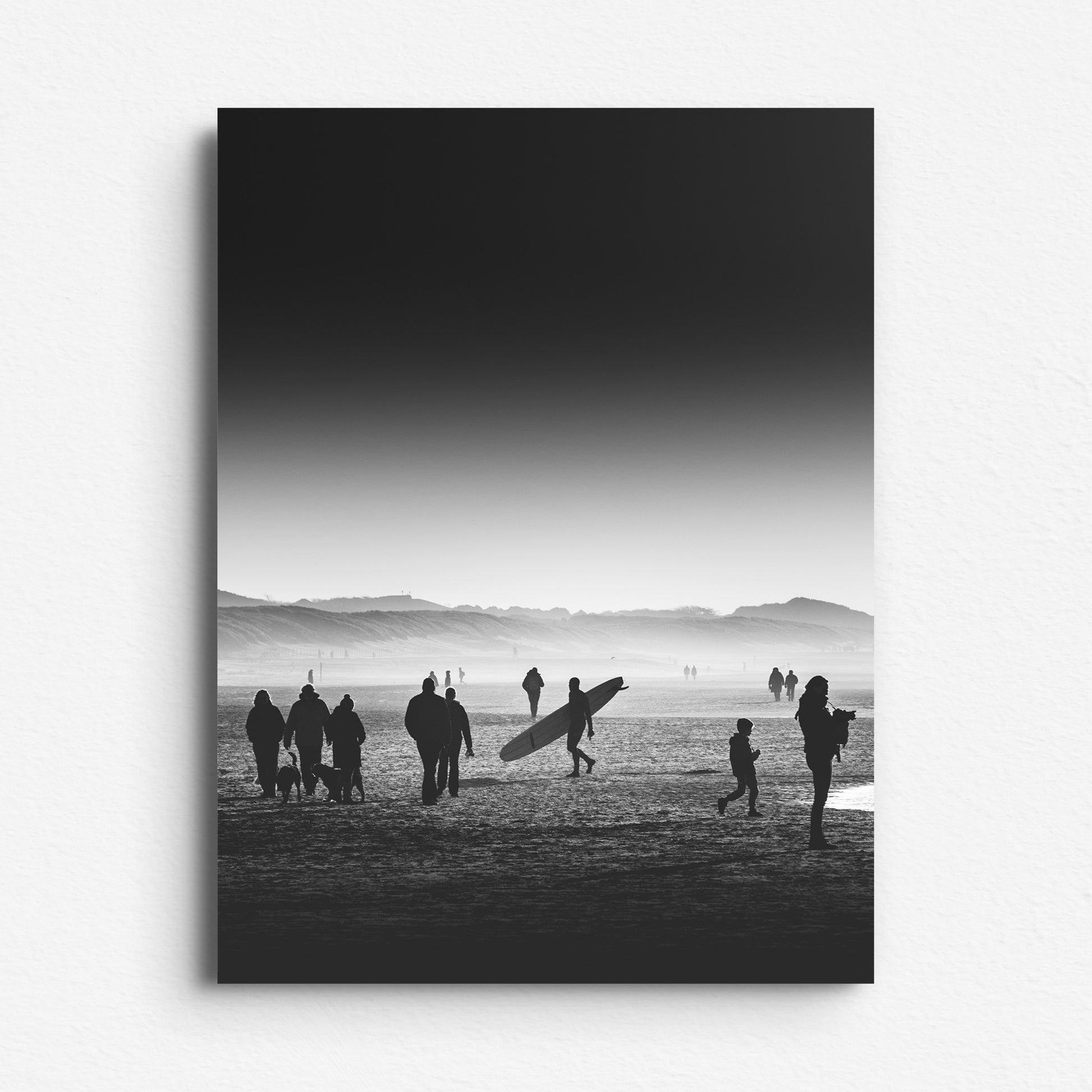 Black and white photo of a man with a longboard walking amid people on the beach. A silhouetted photo. A modern Photo Poster on Aluminum Dibond.