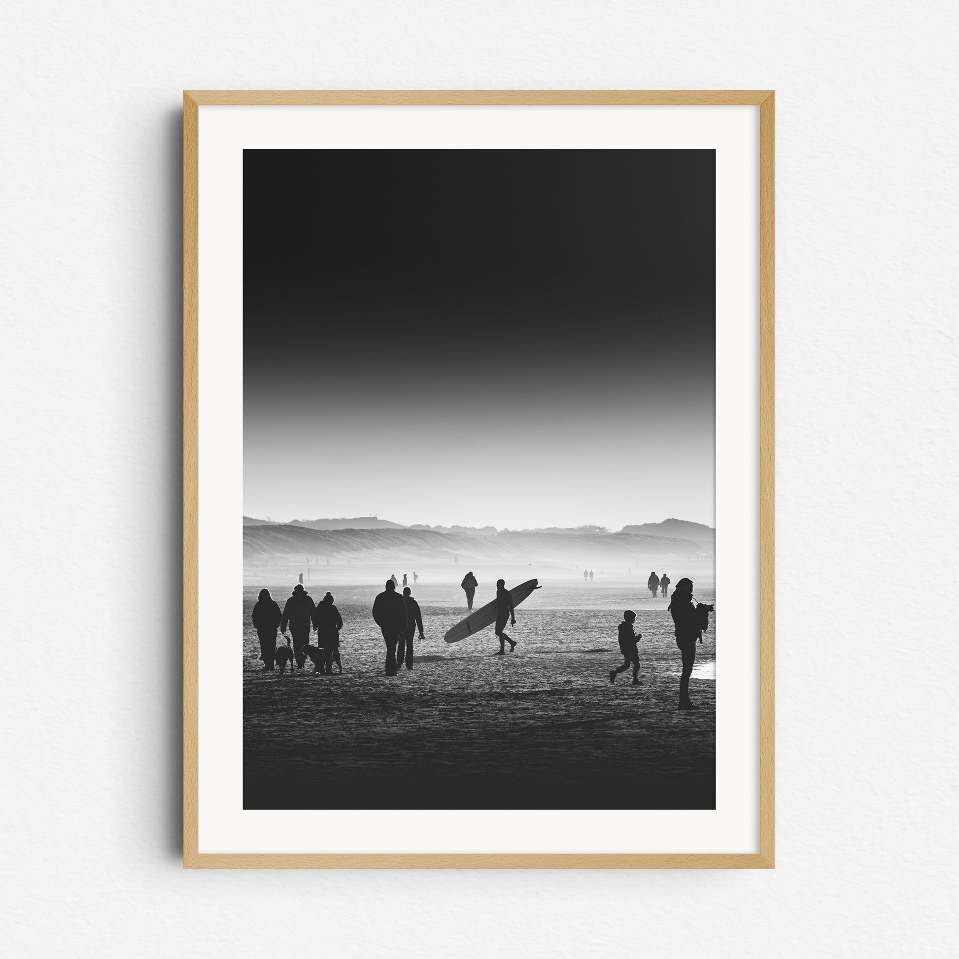 Surfer walking with a longboard on a misty Scheveningen beach in black and white. Dunes in the background. Framed in natural wood, adding a timeless touch to North Sea Surf Photos.