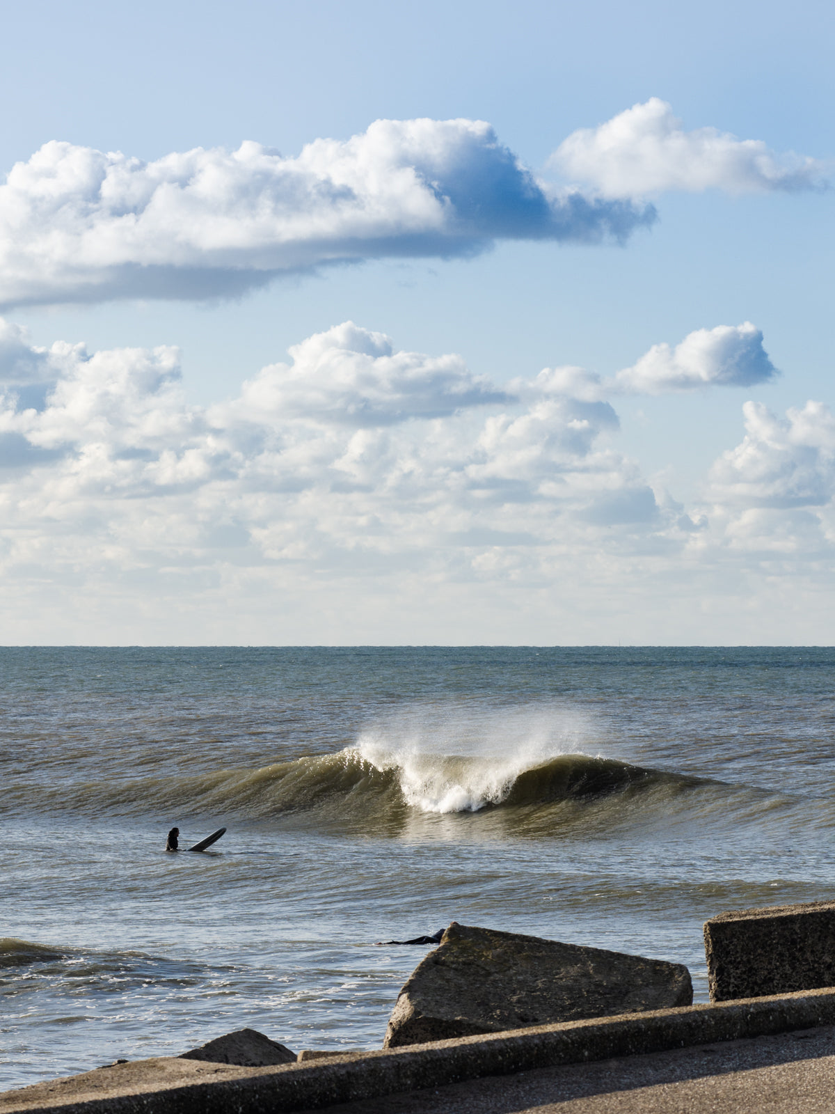 A bright sky with white clouds with a wave breaking. One surfer sits in the sea waiting to catch a wave. Captured by Jop Hermans