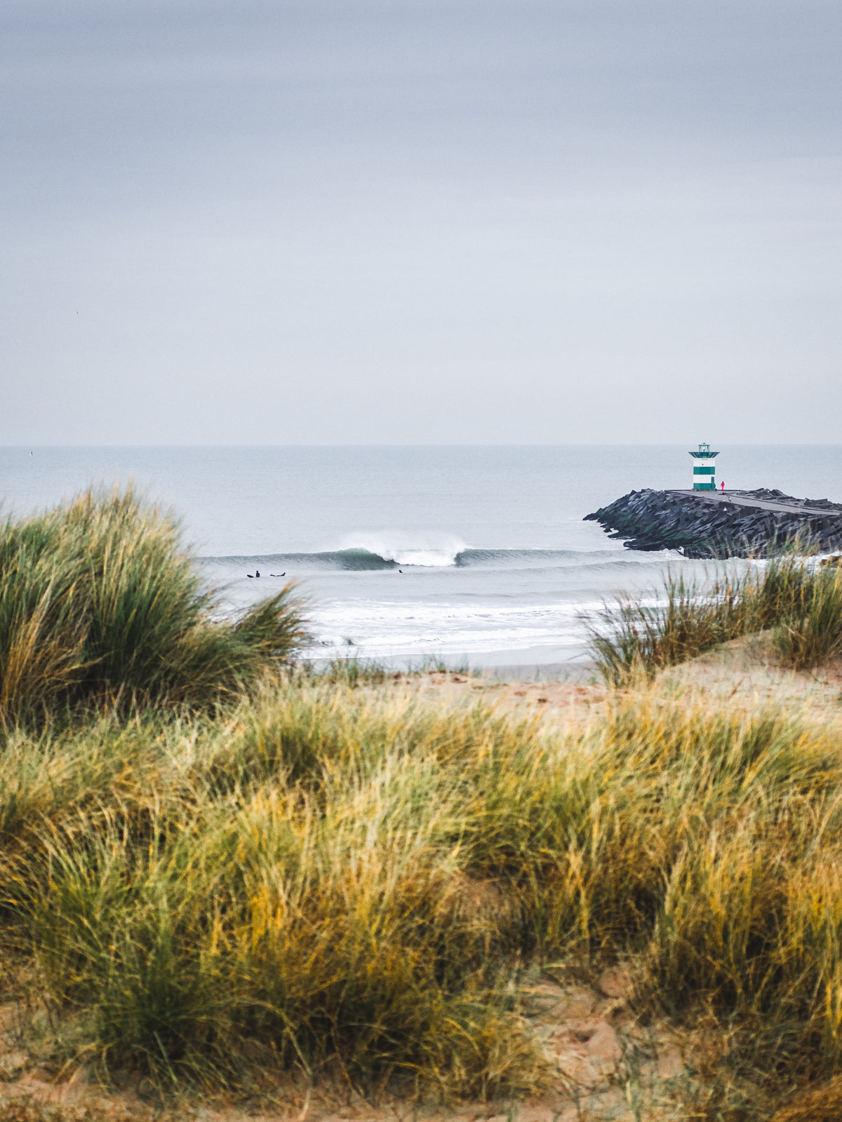 A beautiful breaking wave in The Netherlands shot from an opening in the dunes. A green lighthouse is visible on the Scheveningen harbour breakwall. Photo art from The Hague captured by Jop Hermans.