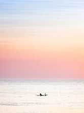 Load image into Gallery viewer, The silhouette of a female surfer paddling in the North sea in Scheveningen, with soft pastel colours in the sky.
