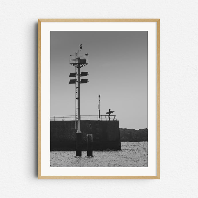 Black and white photo of a Dutch surfer standing in a harbour wall looking out at see. A serene photograph framed in natural wood, captured by Jop Hermans