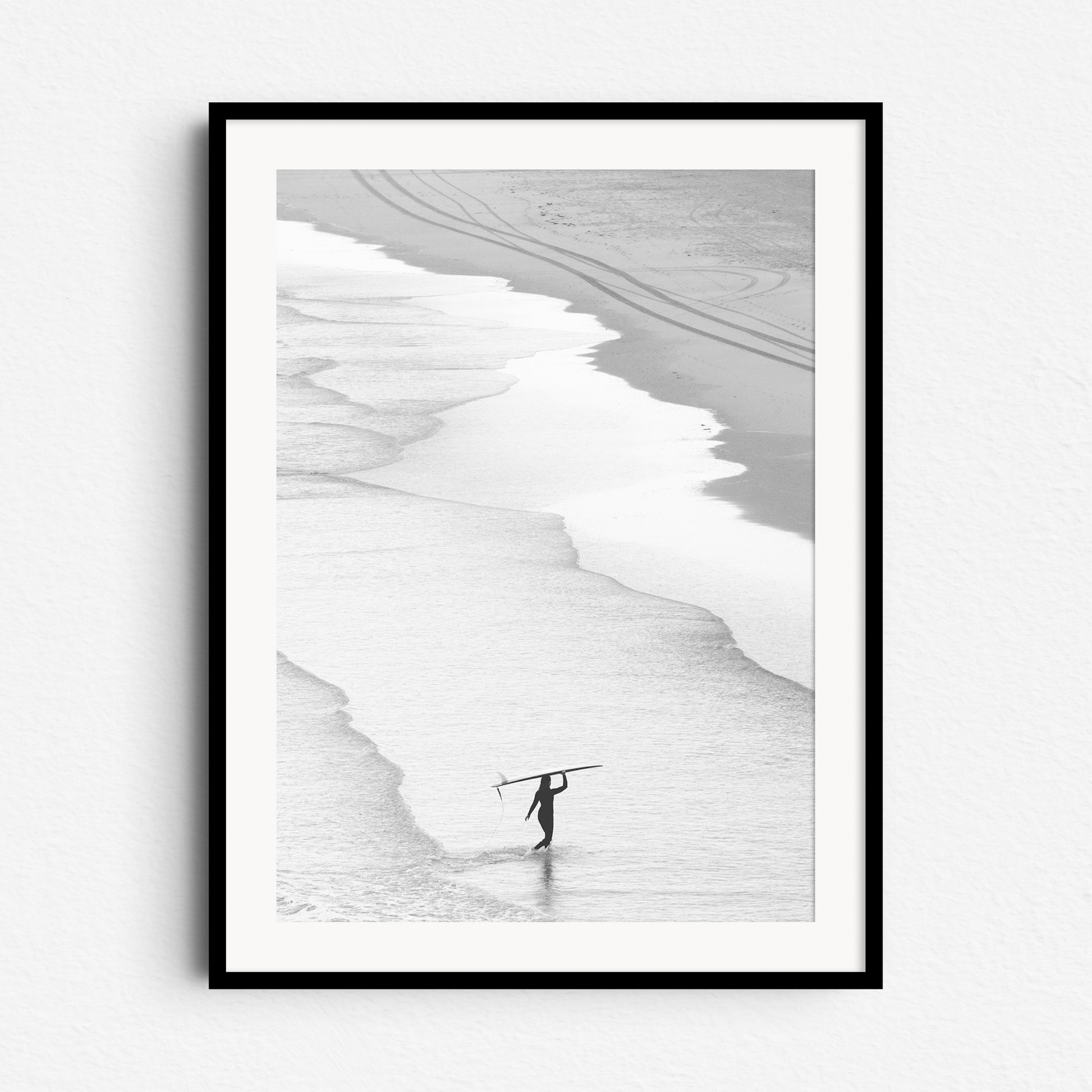 Black and white photo of a woman with a longboard on a Dutch beach, walking through the high tide line. Framed in black wood for a timeless, serene surf scene.