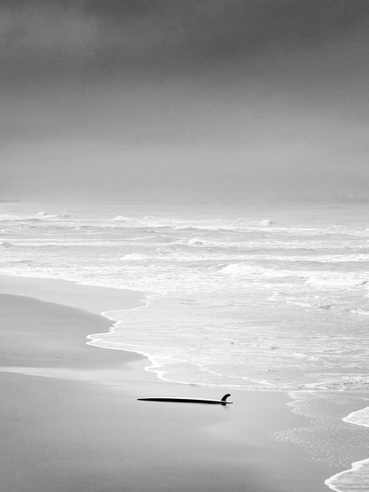 On a deserted Scheveningen beach lays an abandoned surfboard with fin up in the air. The sky looks dark, shot in black and white. Photo art for your wall.