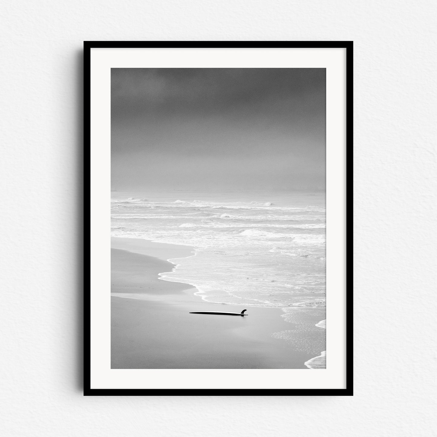 Black and white photo of a surfboard in the sand on a deserted Dutch beach, framed in black wood, ideal for serene Wall decoration.