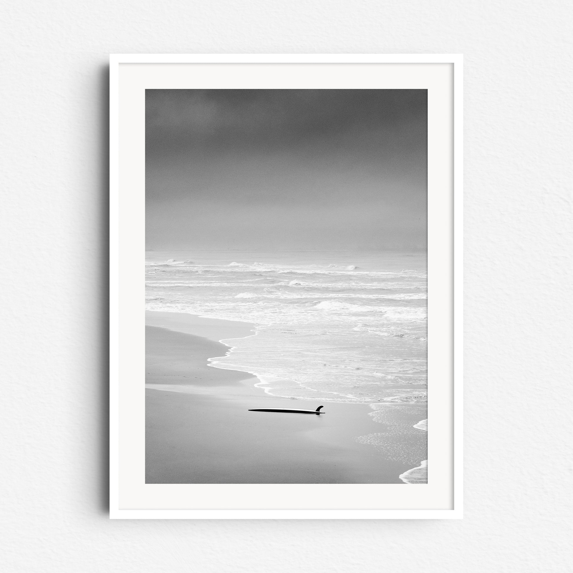 Serene black and white coastal scene in The Hague with a surfboard. The photo poster is framed in white wood, perfect for peaceful North Sea surf lovers.