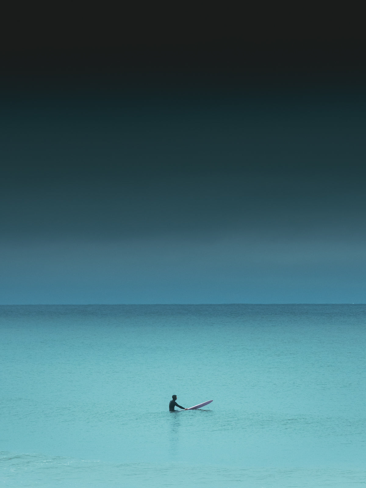 Turquoise coloured sea water with one surfer sitting on a ping surfboard. A photograph with a dark and serene mood. Photo art captured in The Hague.