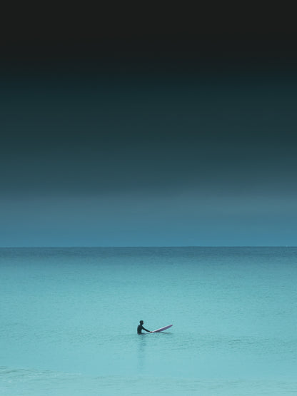 Turquoise coloured sea water with one surfer sitting on a ping surfboard. A photograph with a dark and serene mood. Photo art captured in The Hague.