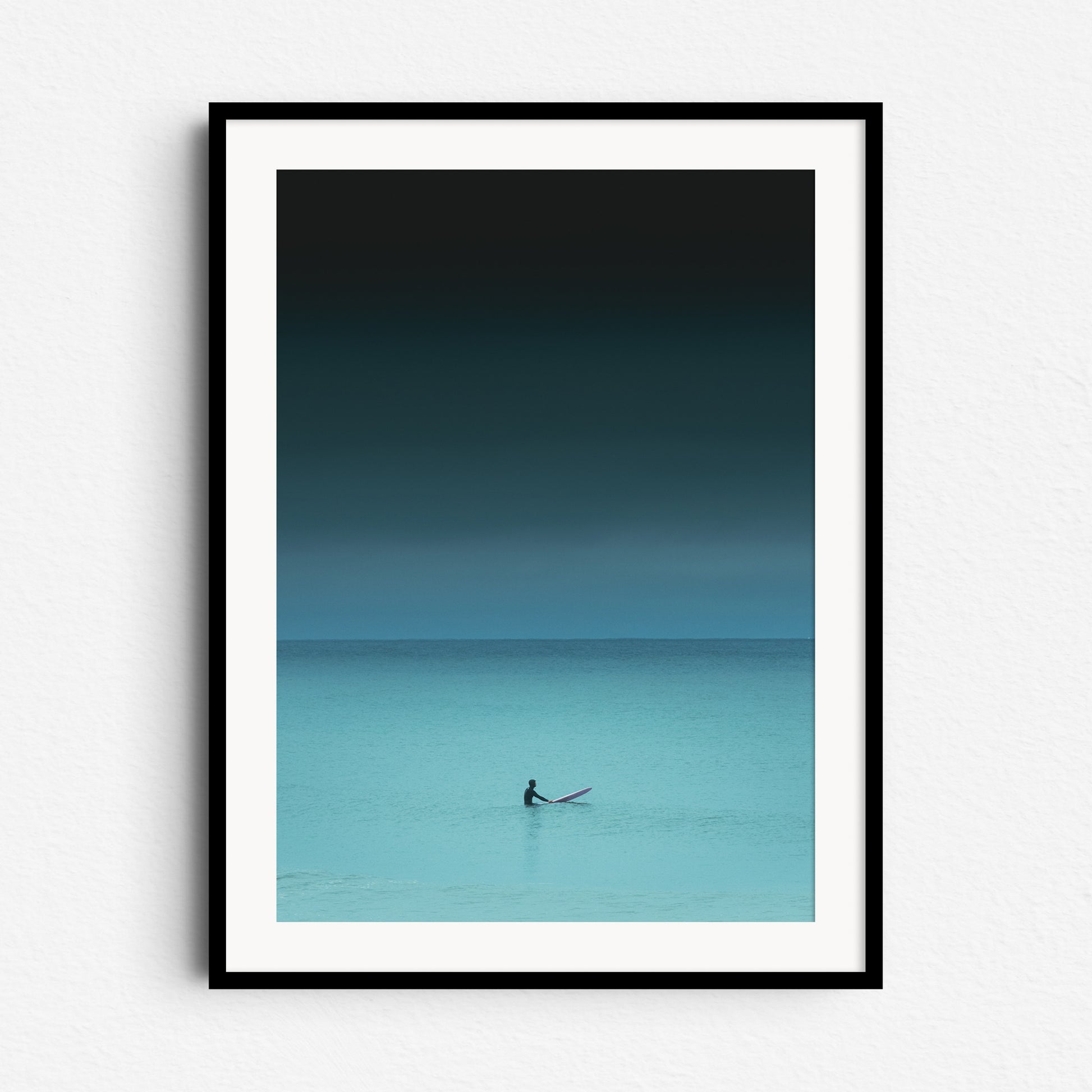 Turquoise photo of a surfer in the North Sea, sitting on a board, framed in black wood. Photo art on the wall for ocean enthusiasts.