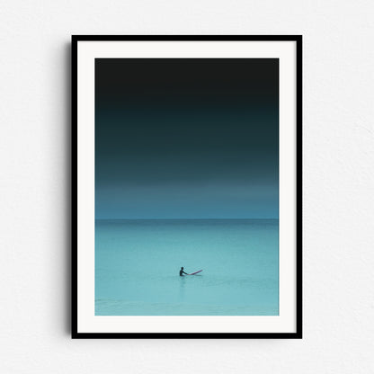 Turquoise photo of a surfer in the North Sea, sitting on a board, framed in black wood. Photo art on the wall for ocean enthusiasts.