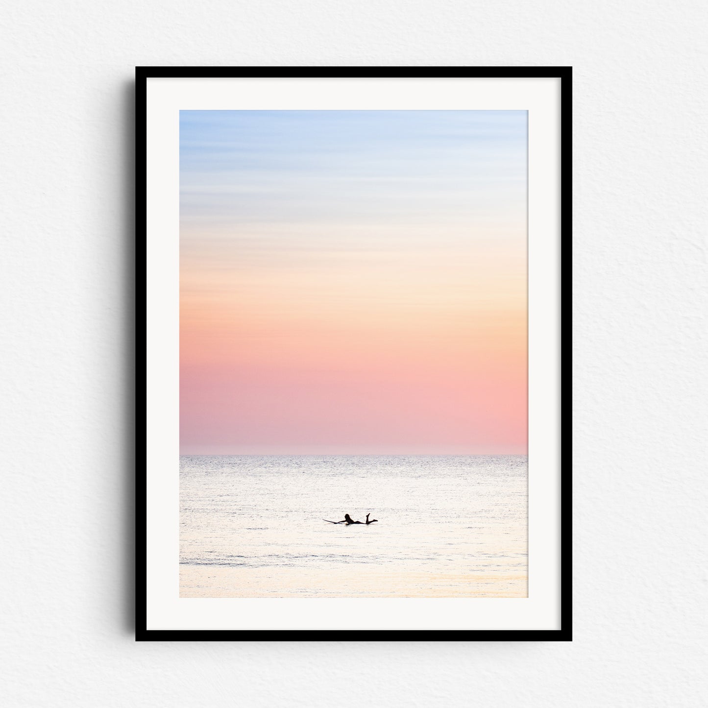 A woman paddles in the North Sea at a summer sunset. A bright, colourful and fresh photo. Framed in black wood to enhance the peaceful pastel glow and serene surf moment.