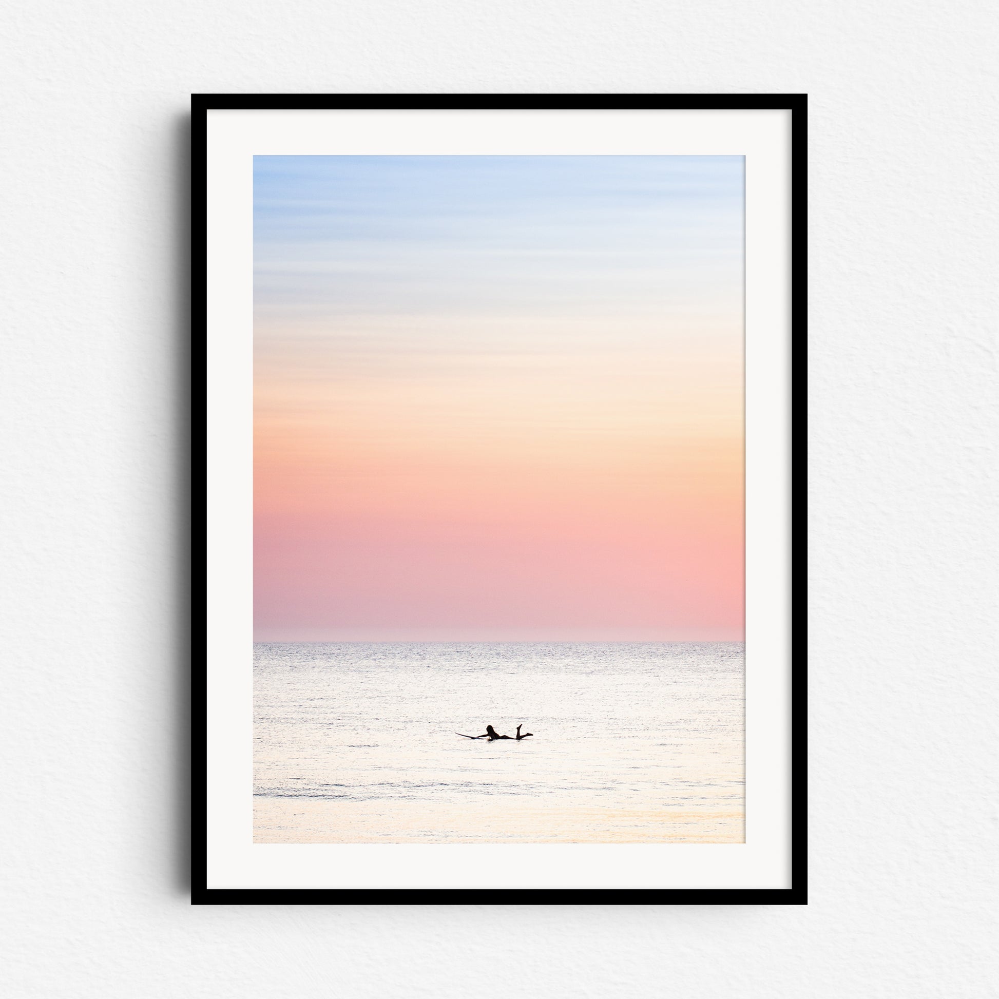 A woman paddles in the North Sea at a summer sunset. A bright, colourful and fresh photo. Framed in black wood to enhance the peaceful pastel glow and serene surf moment.