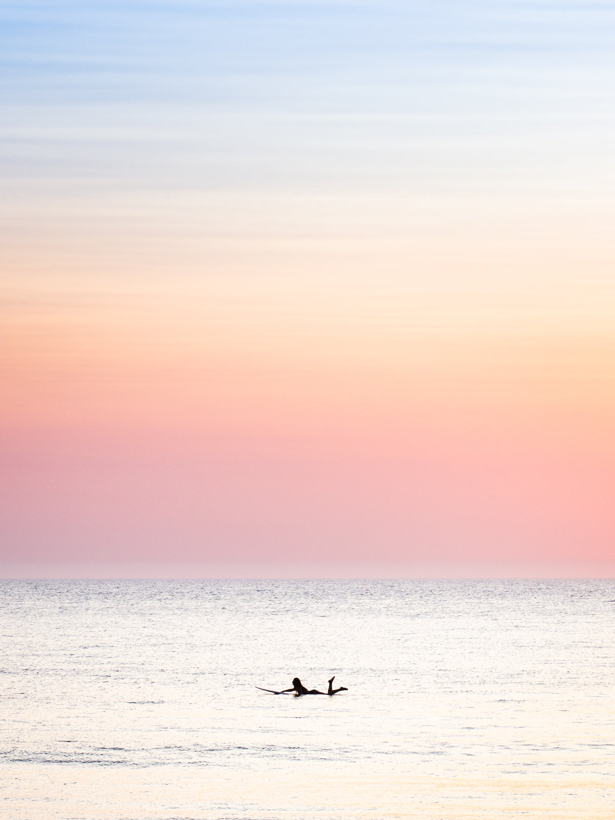 The silhouette of a female surfer paddling in the Dutch North sea, with soft pastel colours in the sky. Surf photography by Jop Hermans