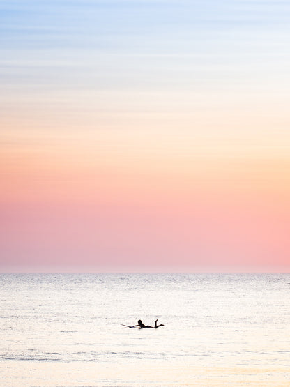 The silhouette of a female surfer paddling in the Dutch North sea, with soft pastel colours in the sky. Surf photography by Jop Hermans