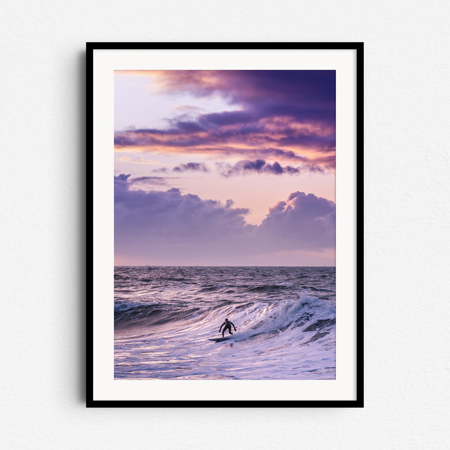 A The Hague surfer rides a wave under purple skies. Framed in black wood, this wall art creates a striking coastal vibe.