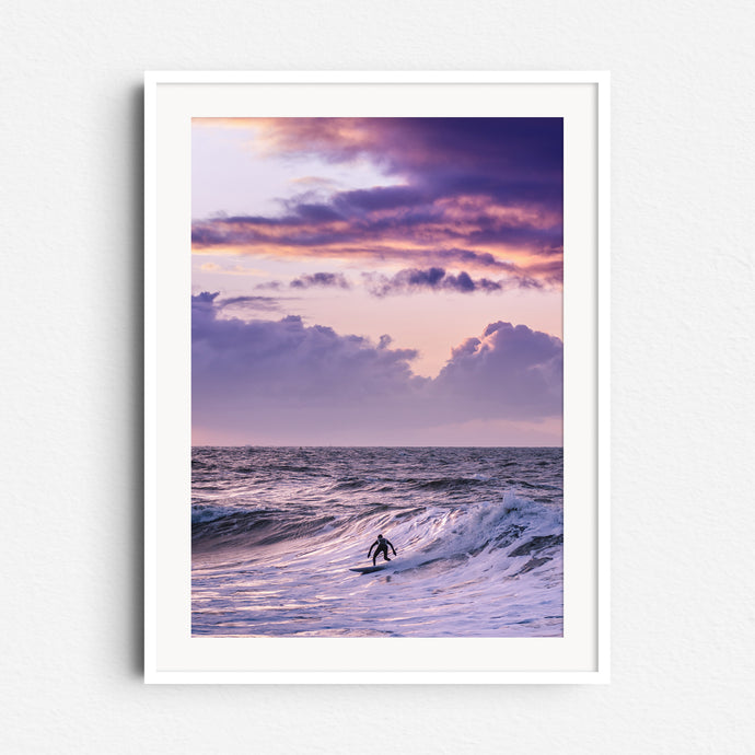 A Scheveningen surfer glides down a wave beneath stunning clouds. Framed in white wood for an elegant, serene addition to your decor.