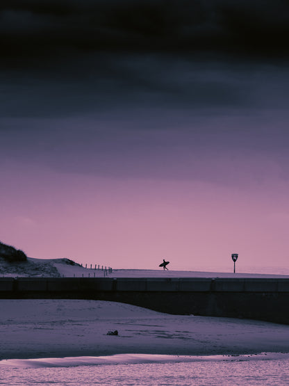 The silhouette of a surfer running across the beach in The Hague, holding a surfboard. The sky is dark and has a purple glow. Classic surf photography from The Netherlands