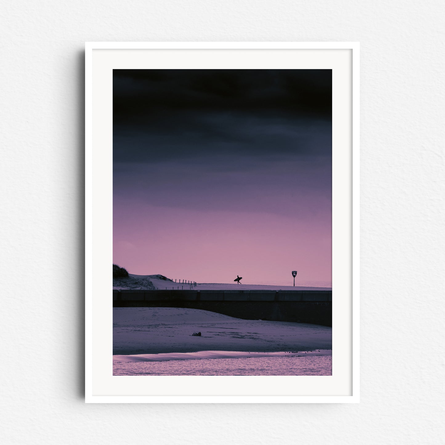 Silhouette of a surfer walking on the south side of Scheveningen harbor during a purple sunset, framed in white wood for a striking and fresh local surf scene.