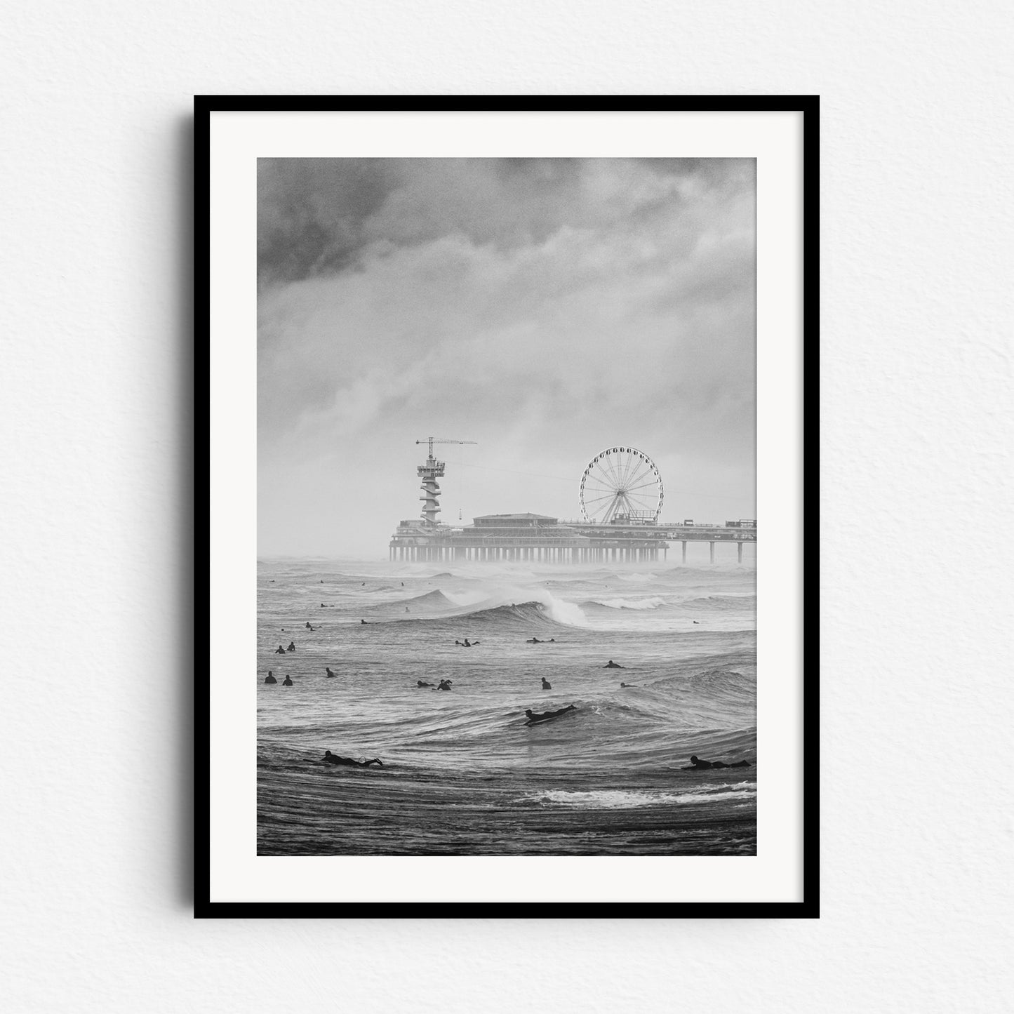 Moody Scheveningen Pier with surfers and beautiful surfing waves. A black and white photo print framed in black wood for a striking, atmospheric wall art piece.