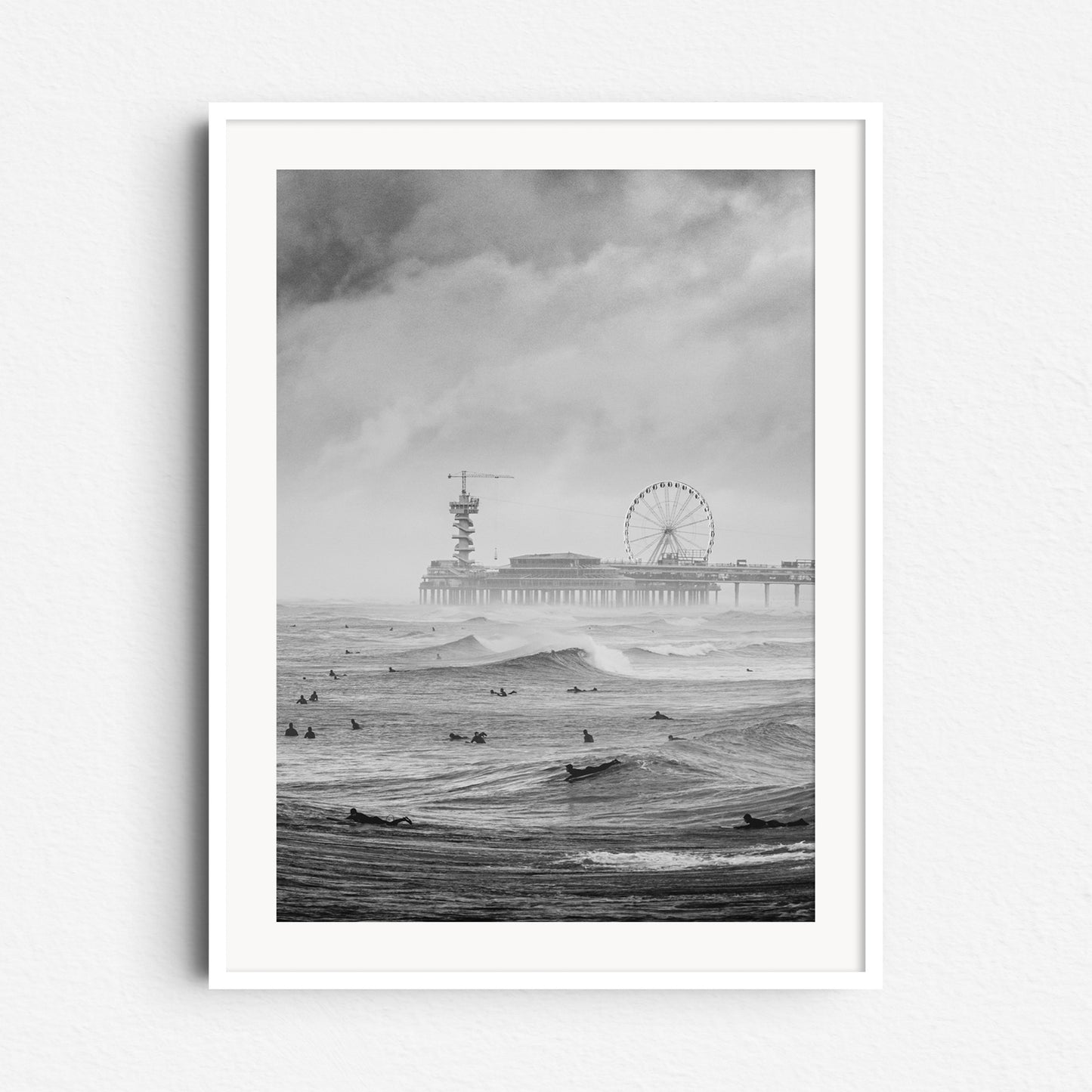 A black and white photograph of North Sea surfers on a grey day, with the Scheveningen pier in the background. Framed in white wood to accentuate the scene’s contrast and mood.