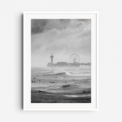 A black and white photograph of North Sea surfers on a grey day, with the Scheveningen pier in the background. Framed in white wood to accentuate the scene’s contrast and mood.