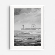 Load image into Gallery viewer, A black and white photograph of North Sea surfers on a grey day, with the Scheveningen pier in the background. Framed in white wood to accentuate the scene’s contrast and mood.
