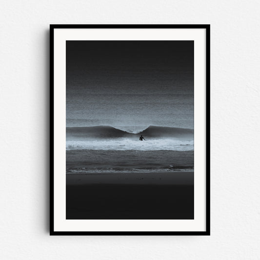 A high-contrast black-and-white photo of a surfer facing a breaking wave. Framed in black wood for bold, dramatic wall art.