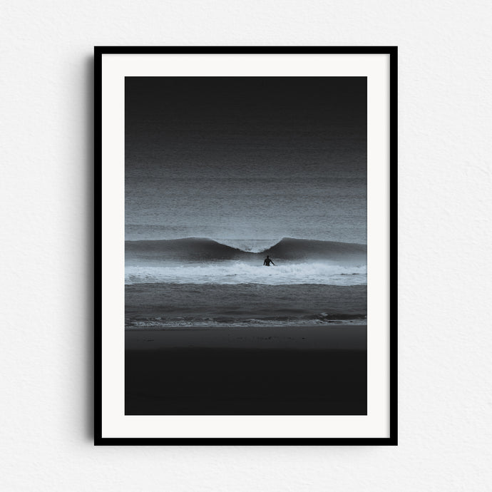 A high-contrast black-and-white photo of a surfer facing a breaking wave. Framed in black wood for bold, dramatic wall art.