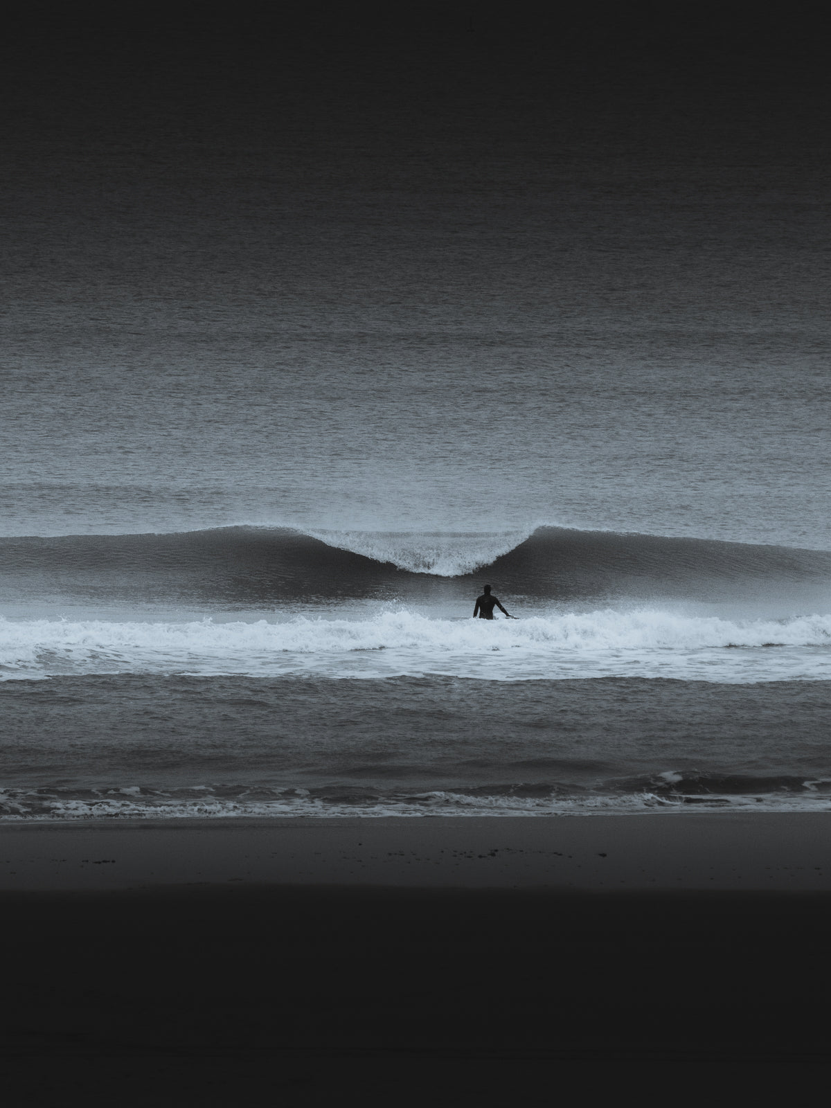 A moody, surreal black-and-white photo of a surfer facing a breaking wave on a beach in Scheveningen. A dramatic wall art piece.