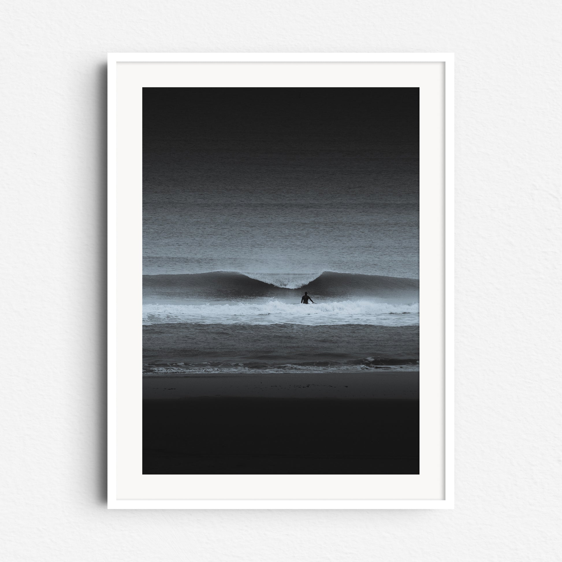 A black-and-white surf scene with a lone surfer and crashing wave. Framed in white wood, ideal for a sleek, minimalist aesthetic.