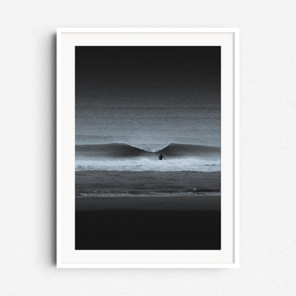 A black-and-white surf scene with a lone surfer and crashing wave. Framed in white wood, ideal for a sleek, minimalist aesthetic.