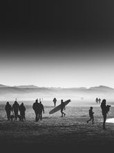 Load image into Gallery viewer, A surfer caries a longboard walking on a misty beach between people, with dunes in the background. Black and white silhouette in Scheveningen.
