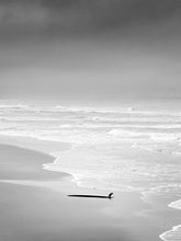 Load image into Gallery viewer, On a deserted Scheveningen beach lays an abandoned surfboard with fin up in the air. The sky looks dark, shot in black and white. 
