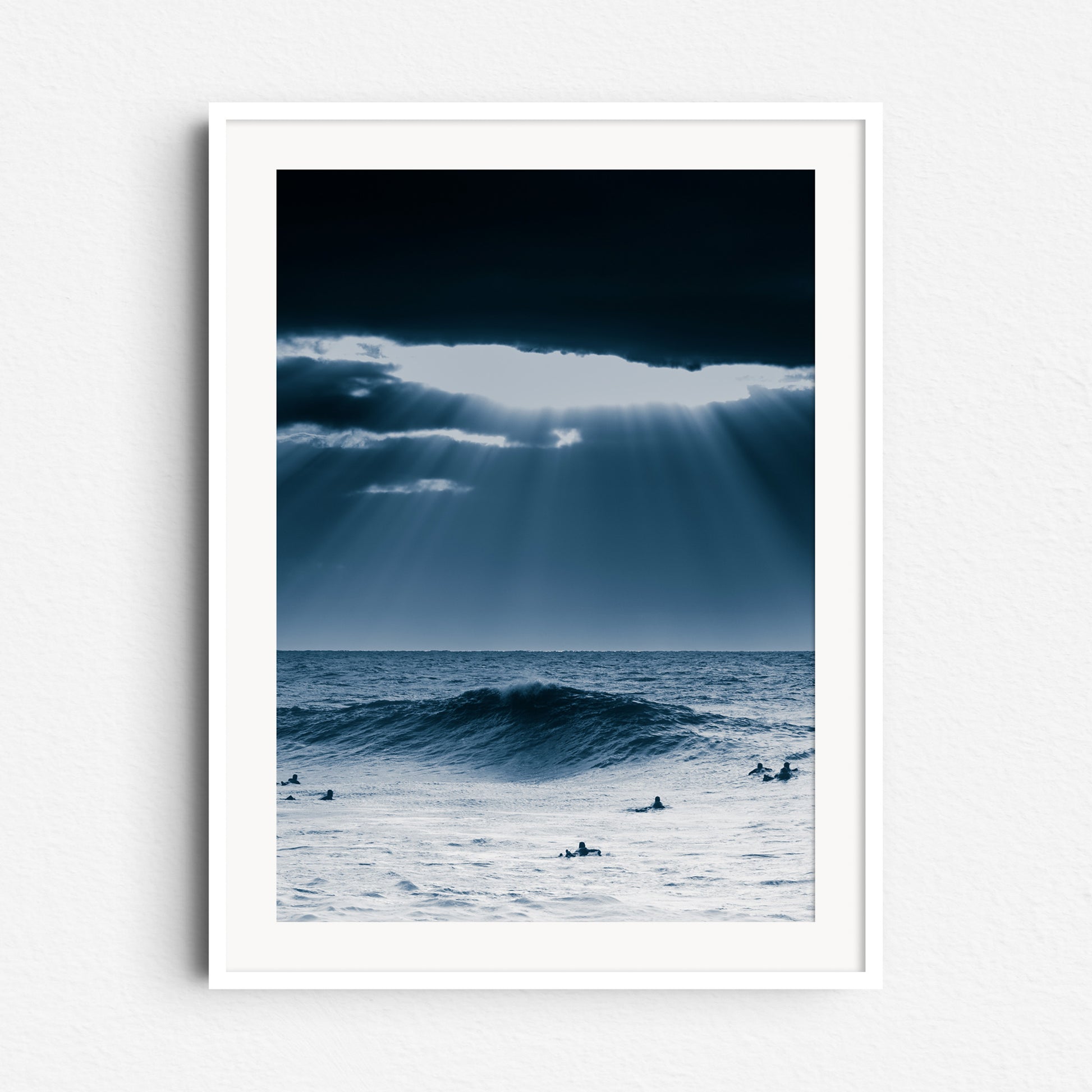 Sunlight bursts through clouds over surfers in the North Sea. Framed in white wood, perfect for a serene and elegant space.