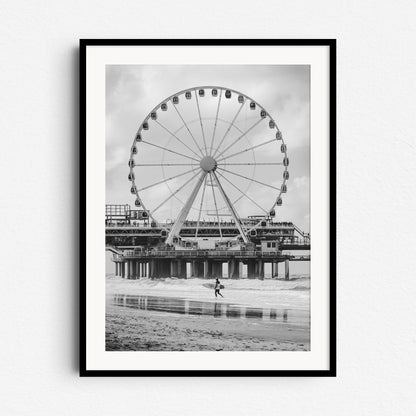 Scheveningen Pier’s ferris wheel with a surfer in front. Black and white North Sea surf photography from The Hague, framed in black wood for a bold and striking wall art piece.